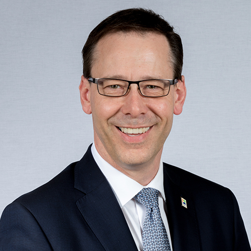 A man in a suit and tie. He's against a white background, and is wearing black glasses.