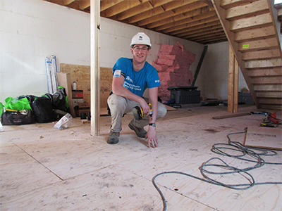 One person smiling at the camera while holding a drill. He appears to be drilling sub floor in a partially finished home.